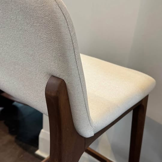 Bar stool with beige cushioned seat and backrest on a wooden frame against a white wall.