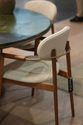 Load image into Gallery viewer, Close-up of a FAZANI dining chair with a warm brown wooden frame, cream fabric seat and back, and a distinctive silver metal loop detail, shown in a real interior setting.