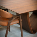 Load image into Gallery viewer, A close-up of a mid-century modern wooden dining table with a sculptural curved base, paired with a brown leather upholstered chair in a softly lit interior.