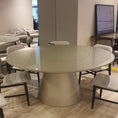 Charger l'image dans la visionneuse de la galerie, A modern round dining table with a glossy light beige top and a tapered metallic base, surrounded by matching upholstered dining chairs in a showroom setting with light wood flooring.