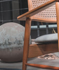 Load image into Gallery viewer, Wooden chair with wicker backrest and gray cushion on a blurred outdoor background