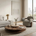 Load image into Gallery viewer, A modern living room scene featuring a round walnut coffee table on a textured light rug, next to a gray upholstered armchair and a light gray sofa with a gold table lamp.