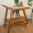 Load image into Gallery viewer, A mid-century modern wooden rolling bar cart with a warm medium-brown finish, styled with glassware and a cocktail shaker on a white tray against a textured white and gray wall.