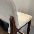 Load image into Gallery viewer, Bar stool with beige cushioned seat and backrest on a wooden frame against a white wall.
