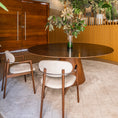 Charger l'image dans la visionneuse de la galerie, A round wooden dining table with a matching pedestal base, surrounded by two mid-century modern wooden chairs with cream upholstered seats, set in a stylish dining room with a crystal chandelier, plants, and a wood cabinet.
