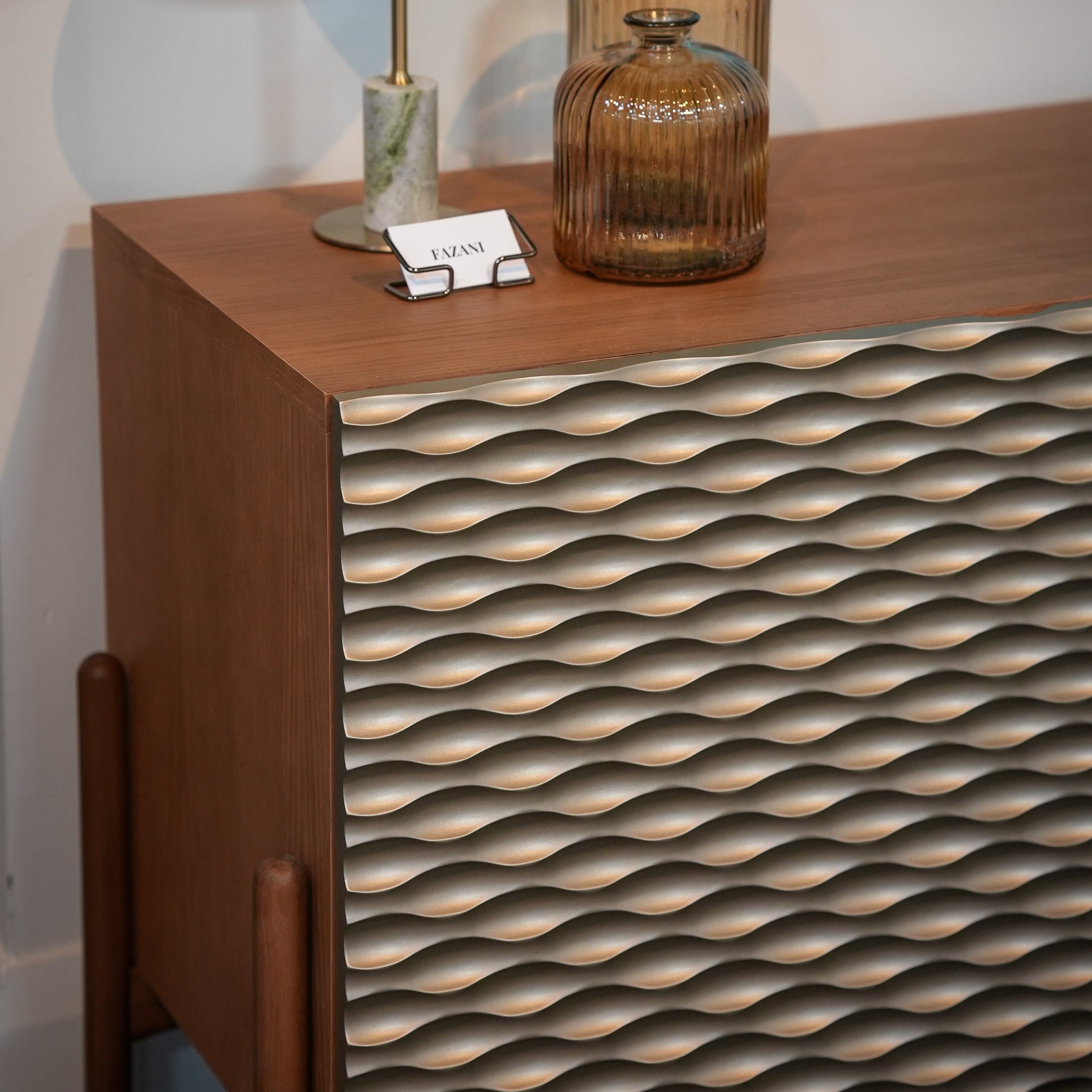 A mid-century modern wooden sideboard or credenza with a wavy textured front panel, styled with a brass table lamp and two amber glass vases against a white wall.