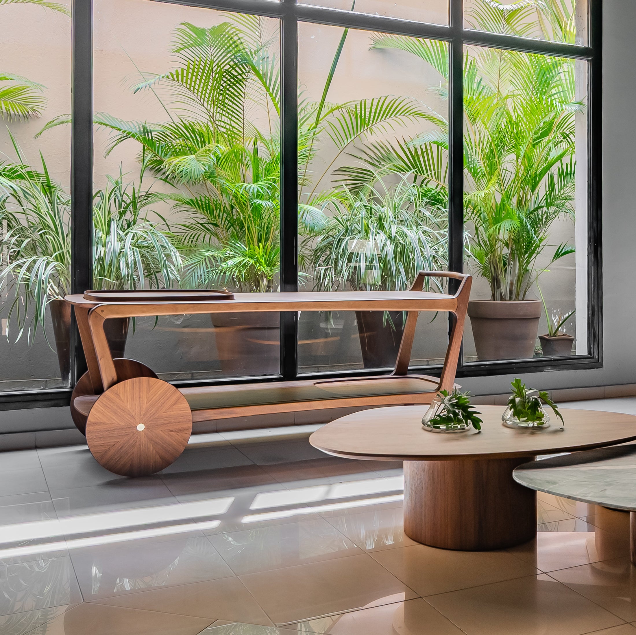 Modern indoor setting with wooden bar cart, tables and plants near large windows.