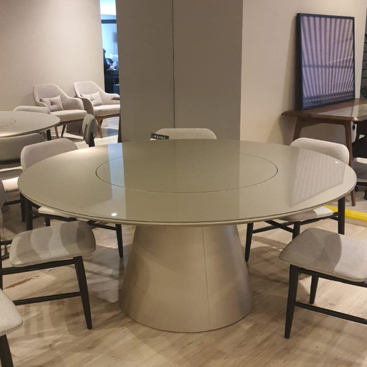 A modern round dining table with a glossy light beige top and a tapered metallic base, surrounded by matching upholstered dining chairs in a showroom setting with light wood flooring.