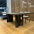 Load image into Gallery viewer, Modern dark brown dining table with a textured wall and chairs in the background
