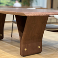 Load image into Gallery viewer, A close-up of a modern wooden dining table with a rich walnut-like finish, featuring a unique sculptural V-shaped base and brass hardware, set in a bright, modern dining room.
