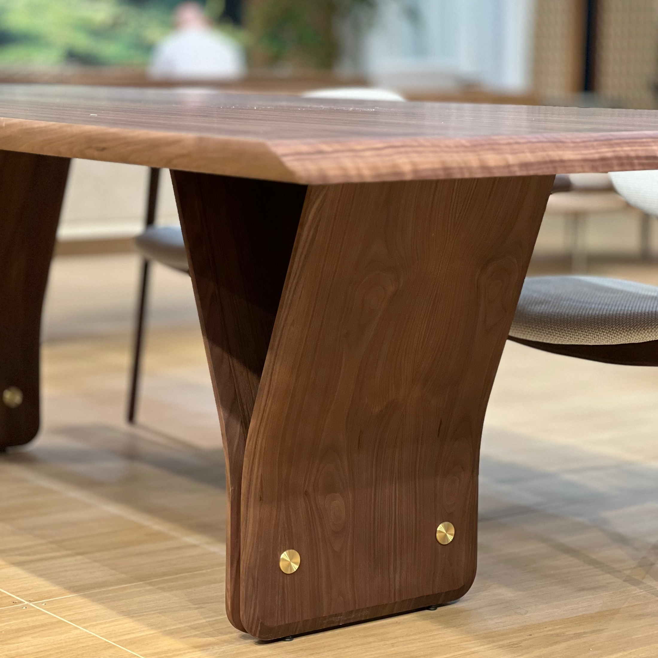 A close-up of a modern wooden dining table with a rich walnut-like finish, featuring a unique sculptural V-shaped base and brass hardware, set in a bright, modern dining room.