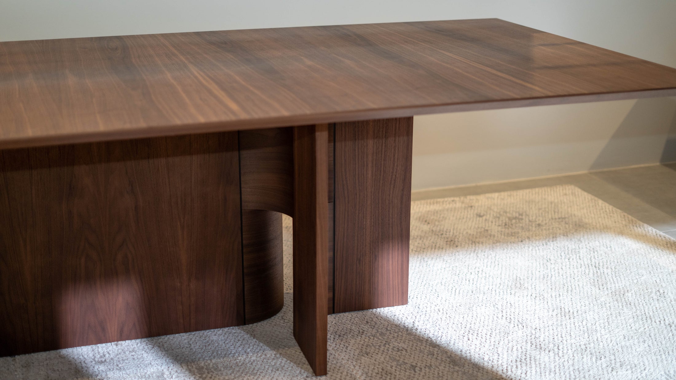 Wooden dining table with a modern design in a room with light-colored carpet and wall.