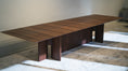 Load image into Gallery viewer, Long wooden dining table in a room with light-colored walls and carpet.
