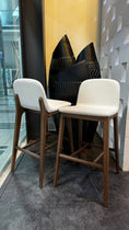 Load image into Gallery viewer, Two white bar stools with wooden legs in front of a decorative wall.
