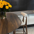 Load image into Gallery viewer, Dining area with a wooden table, chairs, and a vase of yellow roses against a dark wall.
